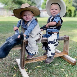 May include: Two young boys dressed in cowboy hats and vests are sitting on a wooden rocking horse. The boy on the left is holding the reins and the boy on the right is holding a toy gun.
