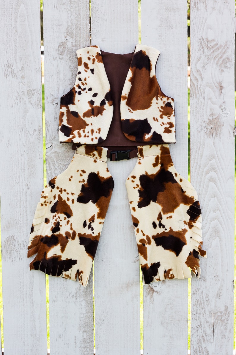 May include: A brown and white cow print vest and chaps with fringe. The vest has a brown lining and a black belt.