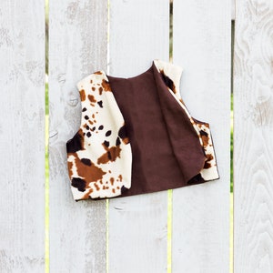 May include: A brown and white cow print vest with a brown lining. The vest is hanging on a white wooden fence.