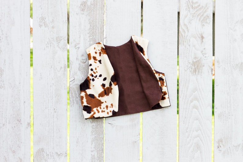 May include: A brown and white cow print vest with a brown lining. The vest is hanging on a white wooden fence.