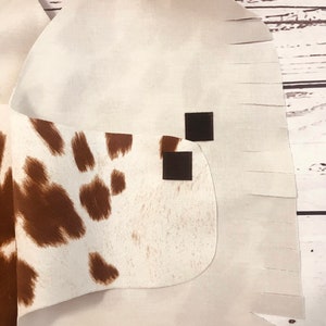 May include: White cowhide vest with brown spots and fringe trim. The vest has two black hook and loop closures.