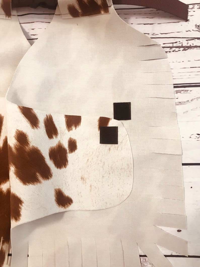 May include: White cowhide vest with brown spots and fringe trim. The vest has two black hook and loop closures.