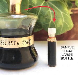 May include: A small sample bottle of Secret de Venus perfume, with a gold cap and a clear glass bottle. The bottle is filled with a dark brown liquid. The sample bottle is next to a larger bottle of the same perfume, with the text "SECRET de VENUS" on the label. The text "SAMPLE FROM LARGE BOTTLE" is written on a white background.