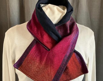May include: A woven scarf with a color gradient from red to purple, with a navy blue collar. The scarf is draped over a white shirt. The scarf's texture is visible, with a geometric pattern.