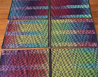 May include: Six rectangular woven placemats in a rainbow of colors, including red, orange, yellow, green, blue, and purple. Each mat has a black border and a textured, patterned design. The mats are arranged on a wooden table.