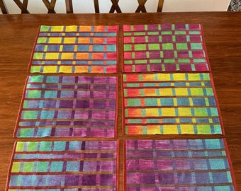 May include: Six colorful woven placemats with a plaid pattern in shades of red, orange, yellow, green, blue, and purple. The placemats are rectangular with a red border and are arranged on a wooden table.