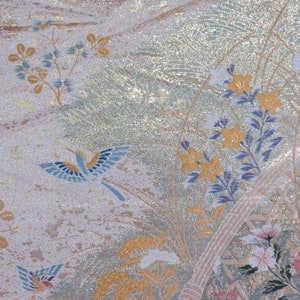 May include: Close-up of a decorative fabric with a shimmering, textured surface. The design features stylized birds in flight, along with floral and botanical elements in shades of gold, blue, pink, and white. The overall effect is elegant and detailed.