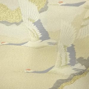May include: A close-up of a fabric with a repeating pattern of flying cranes. The cranes are white with gray and gold accents, set against a pale yellow background. The design has a textured appearance.