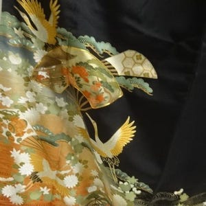 May include: Black fabric with a detailed, colorful design featuring cranes, flowers, and foliage. The artwork includes shades of gold, orange, green, and white, creating a vibrant and elegant aesthetic. The design is likely part of a traditional garment.
