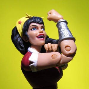 May include: A red, white, and blue Wonder Woman action figure with a red and yellow headband. The figure is posed with her right arm raised in a fist.