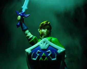 May include: A green and blue plastic action figure of Link from the Legend of Zelda video game series. Link is holding a blue and silver sword and a blue and white shield with the Triforce symbol. The figure is posed in a heroic stance with a determined expression on his face. The background is a dark green with smoke.