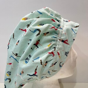 May include: A light blue surgical scrub hat with a pattern of women doing yoga poses in various colors. The hat has a gathered ruffle around the edge.