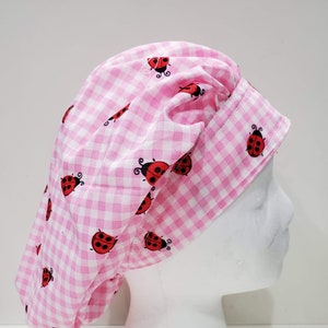Op de afbeelding: Een roze en witte geruite bouffant scrub cap. De cap is versierd met rode lieveheersbeestjes illustraties. De cap is ontworpen om te worden gedragen door medische professionals of in een keuken.