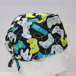 May include: A black scrub cap patterned with white, teal, and lime green video game controllers. The cap is tied at the back and is displayed on a white mannequin head. The controllers are in various orientations.