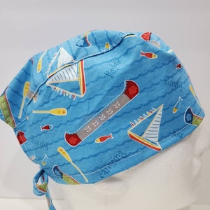 Puede incluir: Un gorro quirúrgico azul con temática náutica. El gorro presenta veleros, peces, remos y las palabras "fishing", "canoeing" y "paddle". El gorro se ata en la parte posterior.