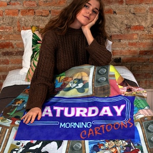 May include: A vibrant blanket displaying cartoon characters and the words "SATURDAY MORNING CARTOONS" in a neon style. The blanket features various cartoon panels and a retro television design.