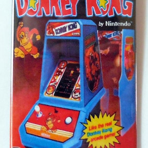 May include: A blue and red Donkey Kong handheld game console with a yellow star and the text "The Official Donkey Kong by Nintendo" on the front. The console is on a red background with the text "Like the real Donkey Kong arcade game!" and "COLECO" on the bottom.