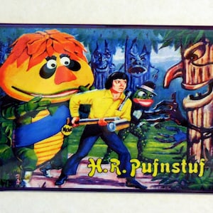 May include: A vintage magnet featuring a cartoon character with orange hair and a yellow body, a boy with a yellow shirt and blue jeans, and a green frog-like creature. The magnet has a purple border and the text "H.R. Pufnstuf" in yellow letters.