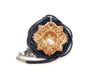 May include: A wooden pendant with a geometric design and a small pearl in the center. The pendant is on a black cord.