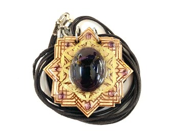 May include: A wooden pendant with a black gemstone in the center, surrounded by a gold-colored mandala design. The pendant is attached to a black cord.