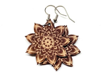 May include: A pair of brown leather earrings with a mandala design. The earrings are made of two layers of leather, with the top layer being a lighter shade of brown than the bottom layer. The earrings are attached to silver hooks.