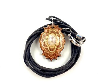 May include: A brown wooden pendant with a gold mandala design and a clear crystal in the center. The pendant is on a black cord with a silver clasp.