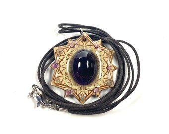 May include: A gold pendant necklace with a large, dark purple gemstone in the center. The pendant is surrounded by a decorative design and small, light purple gemstones. The necklace is made of a dark brown cord.
