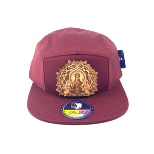 May include: A maroon 5-panel cap with a gold-colored mandala design featuring a seated figure. The cap has a flat brim and a "Pit Bull" logo on the underside of the brim.