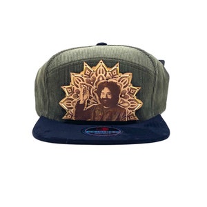 May include: A green corduroy baseball cap with a black brim. The cap features a leather patch with a gold mandala design and a portrait of a man with long hair.