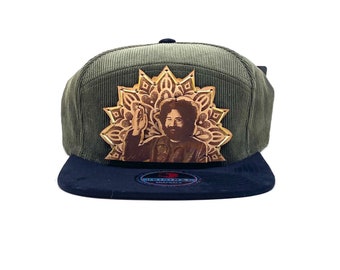 May include: A green corduroy baseball cap with a black brim. The cap features a leather patch with a gold mandala design and a portrait of a man with long hair.