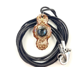 May include: A brown and gold pendant with a geometric design and a dark blue gemstone in the center. The pendant is on a black cord with a silver clasp.