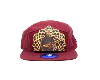 May include: A maroon 5-panel baseball cap with a gold-colored graphic of a musician playing a guitar. The graphic is surrounded by a geometric design.