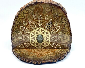 May include: A wooden slice with a geometric design featuring a flower of life pattern and crystals. A gray stone is placed in the center of the design.