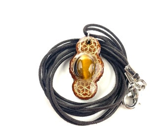 May include: A brown and gold pendant with a tiger's eye stone set in a geometric design. The pendant is on a black cord with a silver clasp.