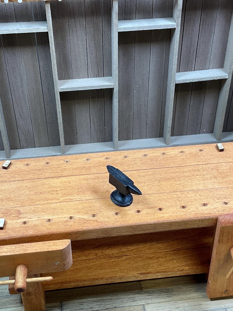 May include: A miniature wooden workbench with a small black metal anvil on top. The workbench has a drawer and a small wooden vise on the side.