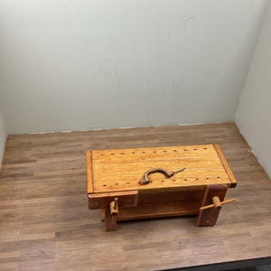 May include: A wooden workbench with a vise and a metal tool on top. The workbench is in a room with wood-look flooring and white walls.