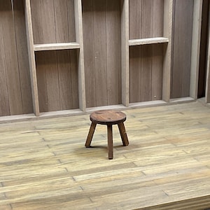 May include: A wooden stool with a round seat and three legs. The stool is made of dark brown wood and has a rustic appearance. The stool is sitting on a wooden floor.