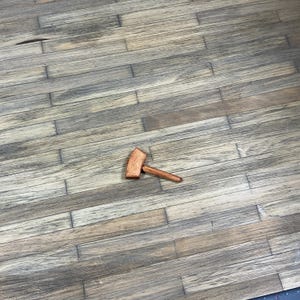 May include: A wooden toy hammer with a brown handle and a light brown head. The hammer is lying on a wooden floor.