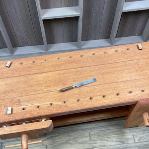 May include: A wooden workbench with a small drawer and a metal chisel on top. The workbench has a light brown finish and is made of solid wood. There are several holes drilled into the top of the workbench, which are likely used for holding tools.
