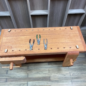May include: Three pairs of pliers on a wooden workbench. The workbench has a shelf and a vise. The pliers are silver with green handles.
