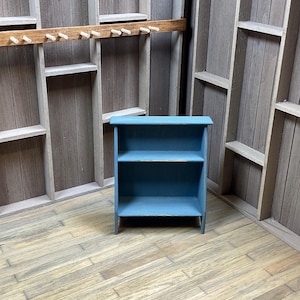 May include: A small, blue wooden shelf with two shelves. The shelf is standing on a wooden floor.