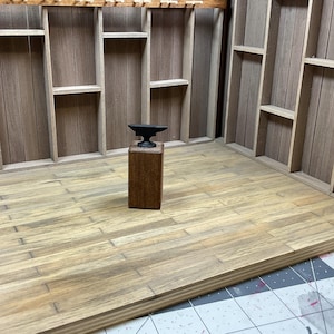 May include: A miniature black metal anvil on a wooden base. The anvil is sitting on a wooden floor in a miniature room with wooden shelves on the walls.