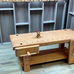 May include: A wooden workbench with a vise and a small red and gold torch on the surface. The workbench has a shelf underneath and is made of natural wood.