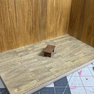 May include: A small wooden stool with a brown finish sits on a light brown wood floor in a miniature room setting.