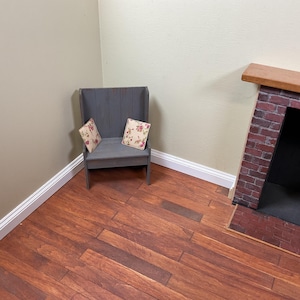 May include: A gray wooden bench with a high back and two floral patterned pillows. The bench is in a corner of a room with wood floors and white baseboards. There is a brick fireplace in the background.