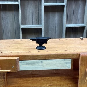 May include: A black metal anvil sits on a wooden workbench with a shelf unit in the background.