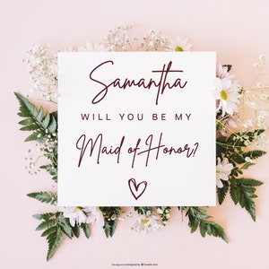 Pode incluir: Um cart&atilde;o branco com o texto "Samantha, voc&ecirc; quer ser minha dama de honra?" em escrita cursiva bord&ocirc; com um pequeno cora&ccedil;&atilde;o desenhado abaixo do texto. O cart&atilde;o est&aacute; cercado por flores brancas e folhas verdes sobre um fundo rosa claro.