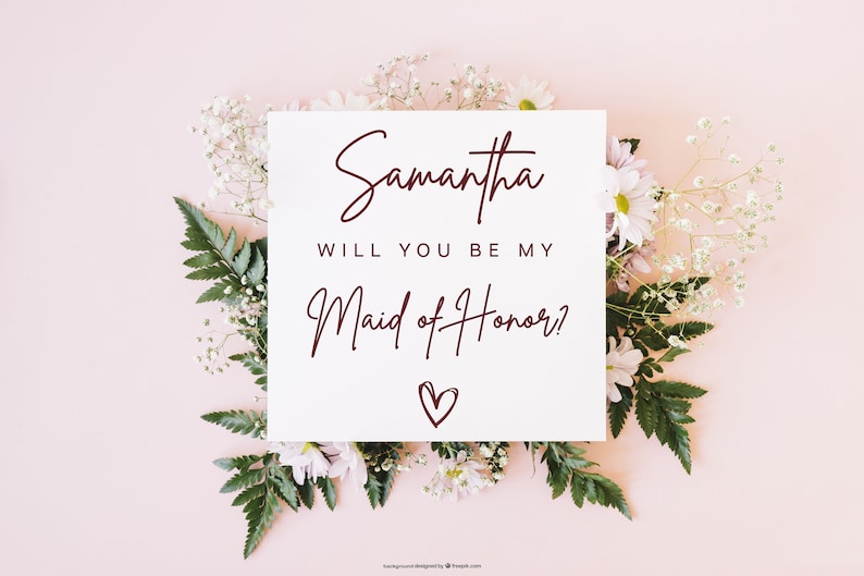 Pode incluir: Um cart&atilde;o branco com o texto "Samantha, voc&ecirc; quer ser minha dama de honra?" em escrita cursiva bord&ocirc; com um pequeno cora&ccedil;&atilde;o desenhado abaixo do texto. O cart&atilde;o est&aacute; cercado por flores brancas e folhas verdes sobre um fundo rosa claro.