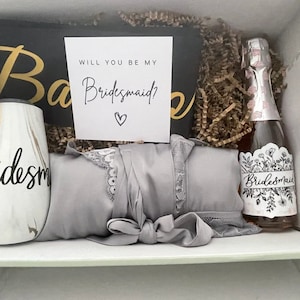Pode incluir: Uma caixa de presente para madrinha de casamento com um roup&atilde;o de cetim cinza, um copo com a palavra "madrinha" nele, uma garrafa de vinho espumante com um r&oacute;tulo floral que diz "madrinha" e um cart&atilde;o que diz "Voc&ecirc; quer ser minha madrinha?"