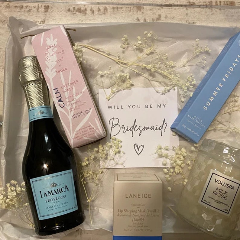 Pode incluir: Uma caixa de presente cheia de itens para uma madrinha de casamento, incluindo uma garrafa de prosecco, uma vela, uma m&aacute;scara labial, um gel de banho e um cart&atilde;o que diz "Voc&ecirc; quer ser minha madrinha?"